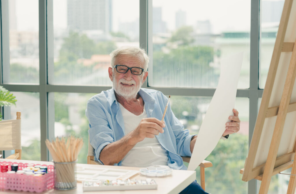 A senior man with glasses holding a small flat brush in his right hand while holding an acrylic paper on his left hand as he smiles directly at the camera.