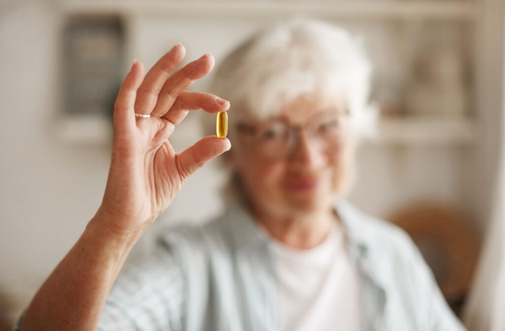 Close-up of an older adult woman with short hair holding up a fish oil pill in her right hand against an orange background