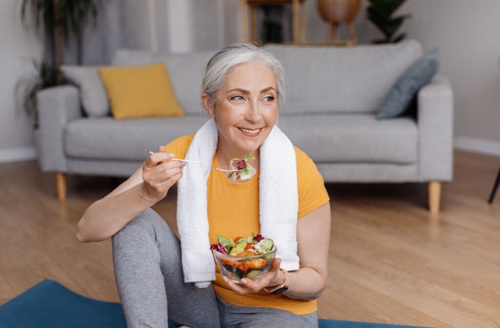 a older woman sitting on a yoga mat after exercise session and eating salad from a bowl