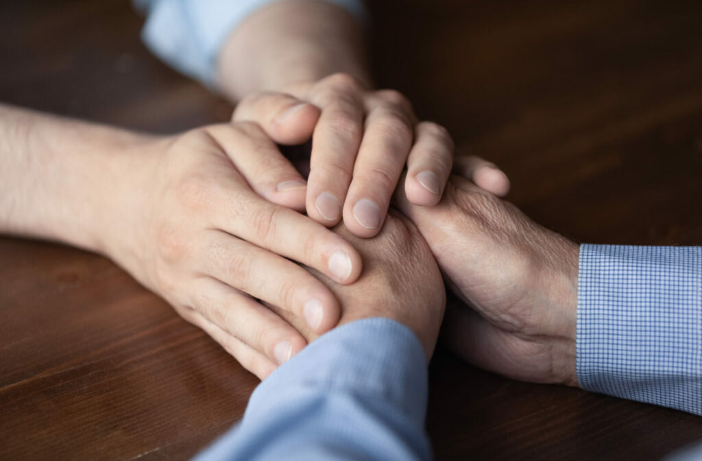 An adult child clasping their older parent's hands in support during a visit in memory care.