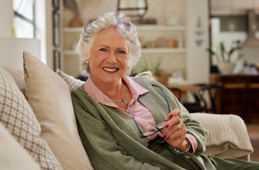 An older adult takes off their glasses and smiles while sitting on a comfortable couch in their home in assisted living